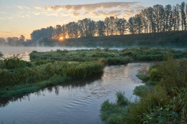 Yaz zamanı. Güneşin doğuşu sisli ve düşünceli bir sis bulutuyla. Sabahın erken saatlerinde sisle kaplı ormanın ve nehrin güzel manzarası. Güneş ışınları..