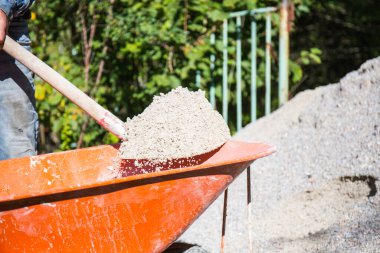 Koyu mavi önlüklü bir işçi, elinde kürekle turuncu bir el arabasıyla çalışan bir inşaat alanının arka planına kum yüklüyor..
