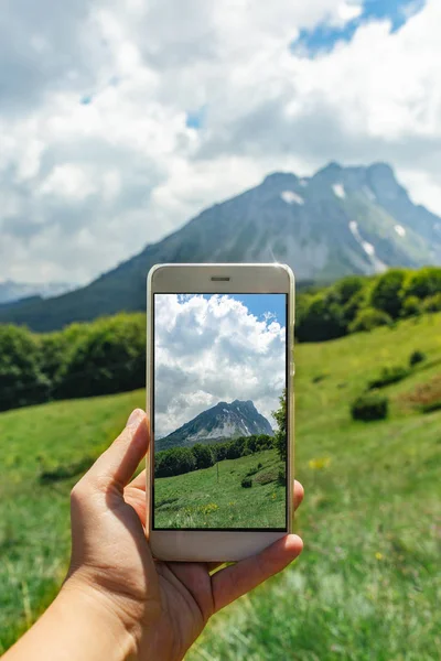 Female hand with smartphone and mountains with green grass and blue sky