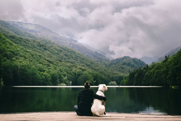 Rear view of woman hugging dog sitting on pier on mountains background