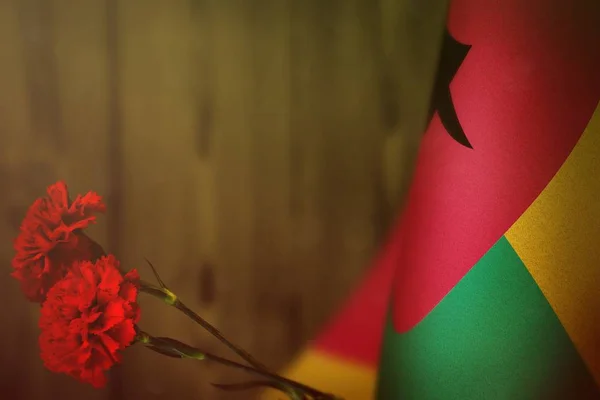 Guinea-Bissau Flagge zu Ehren des Veteranentages oder Gedenktages mit zwei roten Nelkenblumen. Ruhm den Guinea-Bissau-Helden des Kriegskonzepts auf gelber, verschwommener Naturholzwand. Hintergrund. — Stockfoto