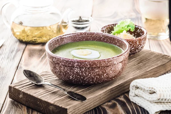 Puree cream soup of green leaf lettuce, spinach and cheese in bowl decorated piece of boiled egg on board for serving dishes on the table, served with green tea in teapot and mug