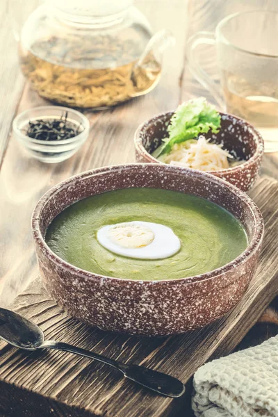 Puree cream soup of green leaf lettuce, spinach and cheese in bowl decorated piece of boiled egg on board for serving dishes on the table, served with green tea in teapot and mug