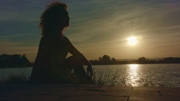 Silhouette giovane donna si siede sulla riva del lago e guarda il tramonto . — Video Stock