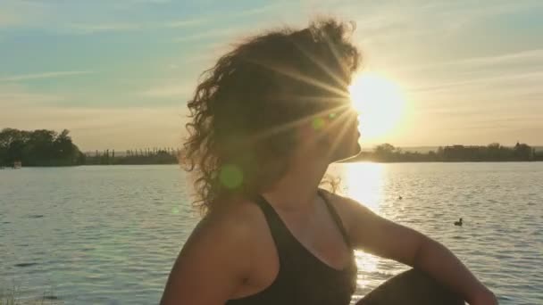 Όμορφη νεαρή γυναίκα κάθεται στην όχθη της λίμνης και φαίνεται στο ηλιοβασίλεμα. — Αρχείο Βίντεο
