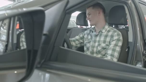 El tipo se sienta en el asiento del conductor. Feliz joven hombre seguro de sí mismo en la luz en el interior, examina el coche del salón — Vídeos de Stock