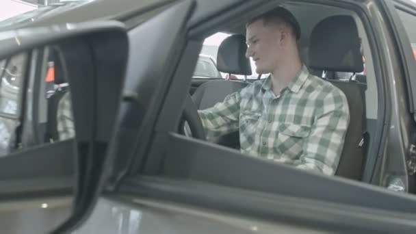 Killen sitter på förarplatsen. Glad ung självsäker man på ljuset i interiören, undersöker salong bilen — Stockvideo