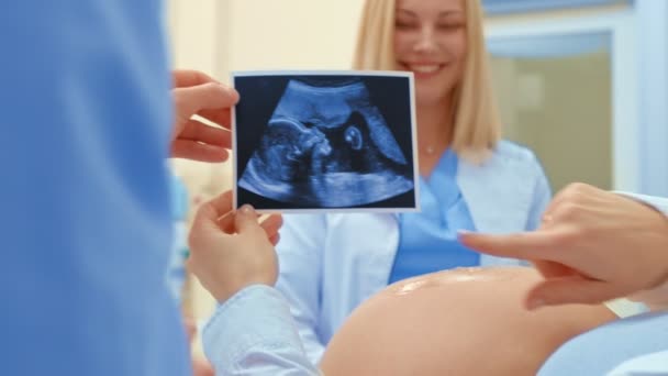 Μια νεαρή έγκυος γυναίκα με τον σύζυγό της φαίνεται στο υπερηχογράφημα το μωρό της. Ένας γιατρός χαμόγελα σε ένα ευτυχισμένο ζευγάρι — Αρχείο Βίντεο