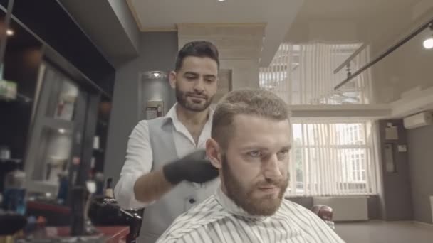 A beautiful hairdresser dressed in a white shirt, gray vest, and black gloves. The young Mister makes a haircut correction. — Stock Video