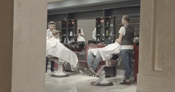 理髪店の椅子に座っている男性のクライアントにサービスを提供するプロの床屋の側からの眺め。髪を切断制服と保護手袋でスタイリッシュな美容師. — ストック動画