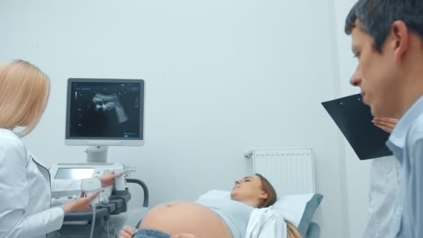 Těhotná žena odpočívá na pohovce v kabinetu ultrazvukové diagnostiky, hovoří a drží ruce s šťastným manželem a ženský doktor na Ultrazvukový test — Stock video