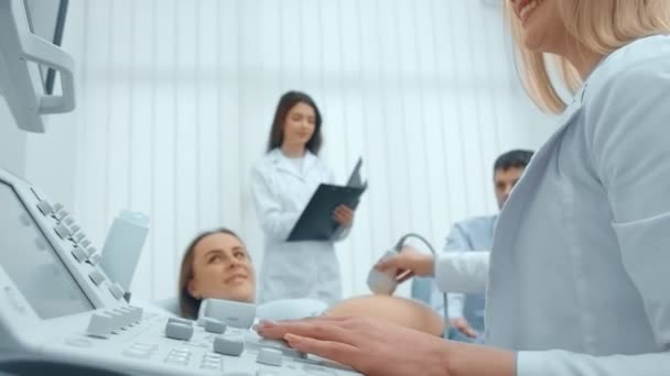 Una donna incinta riposa su un divano in un armadio di diagnosi ecografica, parlare e tenersi per mano con un marito felice e un medico donna per fare test ecografici — Video Stock