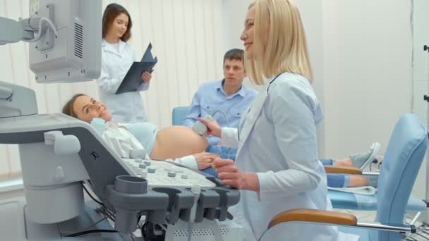 Lekarz wykonuje badanie ultrasonograficzne dla kobiety w ciąży w szpitalu. Szczęśliwy człowiek trzymając żonę ręką i oglądając ekran — Wideo stockowe