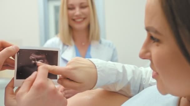 Médico femenino mostrando la ecografía de la mujer embarazada en la foto explicándolo y sonriendo — Vídeo de stock