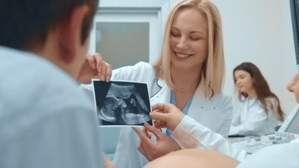 Médico femenino mostrando la ecografía de la mujer embarazada en la foto explicándolo y sonriendo — Vídeo de stock