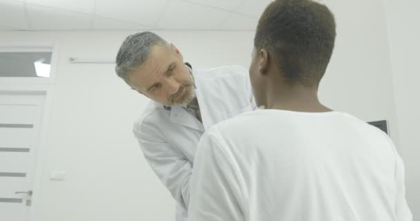 De senior arts onderzoekt de patiënt op het cervicale gebied. Een Afro-Amerikaan zit op een bank en wacht op een specialisten resultaat. — Stockvideo