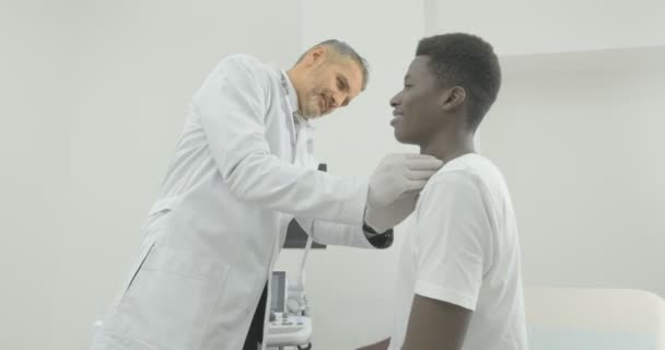 El médico senior examina al paciente en el área cervical. Joven afroamericano se sienta en el sofá y espera el resultado de un especialista . — Vídeo de stock