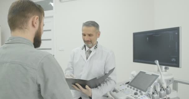 Junger Mann im grauen Hemd auf Absprache mit seinem Therapeuten. Patient schaut Arzt an, hört zu. Reifer Arzt in weißer Arztuniform blickt nach unten. — Stockvideo