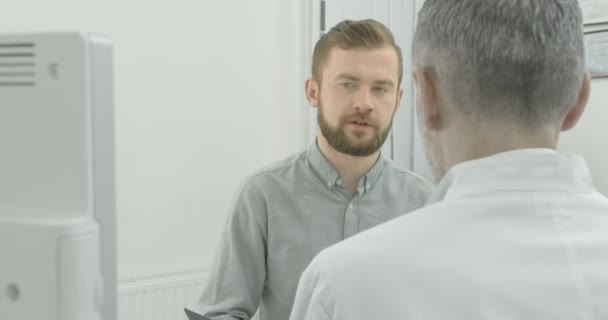 Junger Mann im grauen Hemd auf Absprache mit seinem Therapeuten. Patient schaut Arzt an, hört zu. Reifer Arzt in weißer Arztuniform blickt nach unten. — Stockvideo