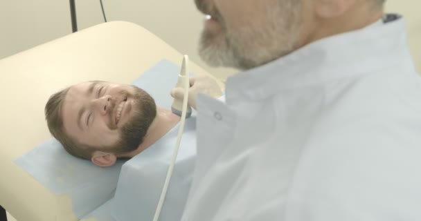 Вид збоку бородатого чоловіка лежить на огляді в медичному кабінеті. Доктор в медичній білій рукавичці з використанням ультразвукового зонда для обстеження . — стокове відео