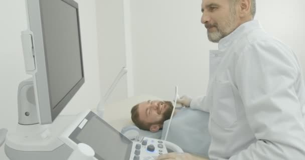 Серьезный доктор, сидящий с клиентом в медицинском кабинете. Терапевт делает ультразвуковую диагностику, смотрит на монитор, держит зонд. Специалист по изучению лимфоузлов на шее . — стоковое видео