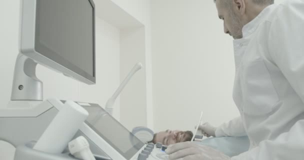 Médecin sérieux assis avec le client dans l'armoire médicale. Thérapeute faisant des diagnostics par ultrasons, regardant le moniteur, tenant la sonde. Spécialiste examinant les ganglions lymphatiques sur le cou . — Video