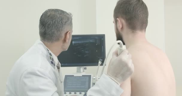 Arts en patiënt zitten, kijkend naar het scherm van de monitor, terwijl de arts te doen scan van Mans schouder. Therapeut met behulp van echografie sonde, doen diagnose met speciale apparatuur. — Stockvideo