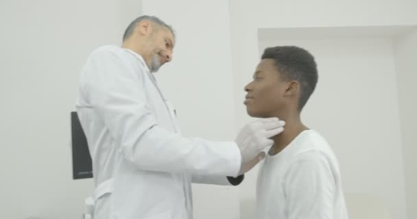 Afrikanischer junger Mann im medizinischen Kabinett. Arzt in weißem Arztkittel und Handschuhen, der mit dem Klienten in der Klinik arbeitet. Arzt tastet Lymphknoten mit Fingern ab — Stockvideo
