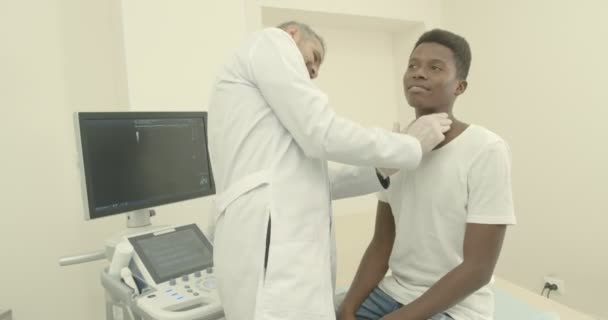 Joven africano en el gabinete médico. Doctor vistiendo bata médica blanca y guantes, trabajando con el cliente en la clínica. Médico palpando ganglio linfático con los dedos — Vídeos de Stock