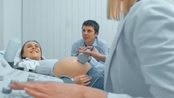 Беременная женщина и ее муж смотрят на ультразвуковое сканирование ребенка. Муж держит жену за руку. — стоковое видео