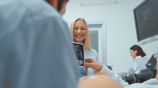 Un jeune couple regardant une image de balayages ultrasonores d'un enfant sur le canapé. Docteur femme sourit avec un couple heureux — Video