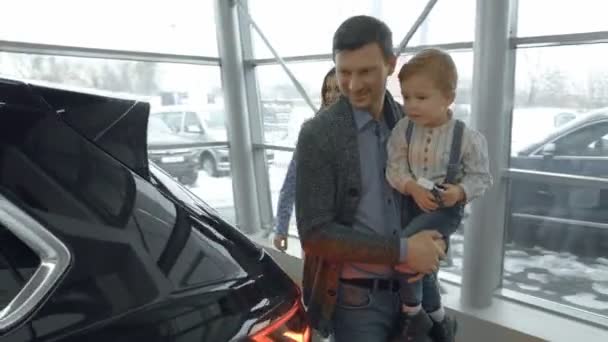 Obserwując nowe, drogie czarny samochód w nowoczesny showroom szczęśliwą rodzinę. Ojciec posiadania dzieciaka z strony, otwieranie bagażnika samochodu, rodzice, stojąc z dziećmi, uśmiechając się, patrząc na samochód. — Wideo stockowe