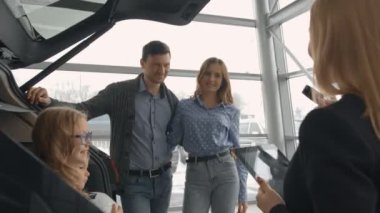 Genç bir çift kameraya bakarken, gülümseyerek, yeni bir arabanın bagajında oturan çocuklar. Yönetici, potansiyel araba alıcılar ile bir bayilik kabinçalışan güzel bir kadın, elinde bir klasör tutar