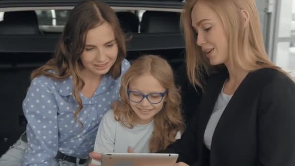 Kızıyla birlikte araba bagajında oturan genç bir kadın araba galerisinden yeni bir otomobil alıyor. Güzel kadın, araba satıcısı elinde belgeyle siyah dosya tutuyor.. — Stok video