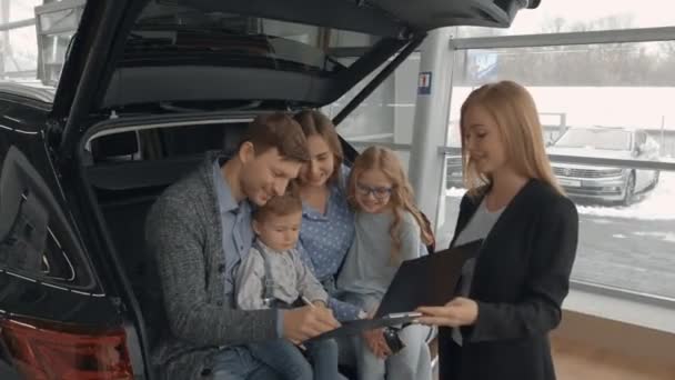 Familia feliz sentado en el maletero del coche, la compra de automóviles nuevos en la concesionaria de automóviles. Hermosa mujer, concesionario de coches sosteniendo carpeta negra con documento. Hombre contrato de firma, compra de vehículo . — Vídeos de Stock