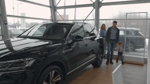 Familia observando el nuevo automóvil negro caro, caminando en la sala de exposición con grandes ventanales de concesionario de automóviles modernos. Hermosa madre e hija cogidas de la mano, mirando el coche . — Vídeo de stock