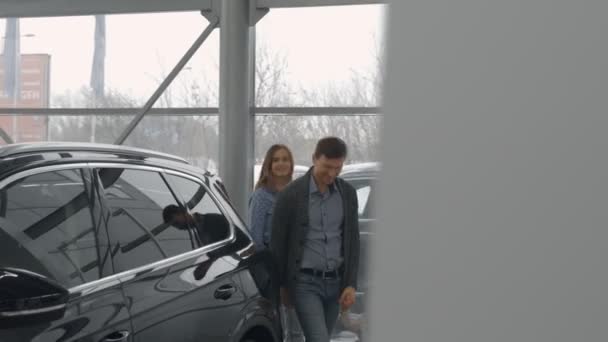 Familia observando el nuevo automóvil negro caro, caminando en la sala de exposición con grandes ventanales de concesionario de automóviles modernos. Hermosa madre e hija cogidas de la mano, mirando el coche . — Vídeo de stock