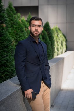 Kendine güvenen genç businessmanin mavi bir takım elbise ayakta şehirde kullanarak. İş adamı olan bir fren ve ofis dışında kamera bakıyor.