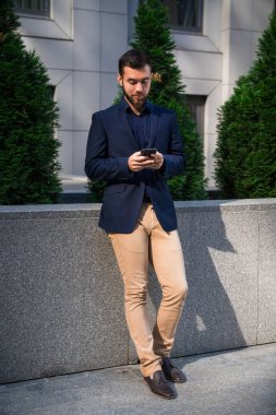 Kendine güvenen genç businessmanin mavi bir takım elbise şehirde cep telefonu kullanarak. Adam manifatura bir mesaj outddor ofis.