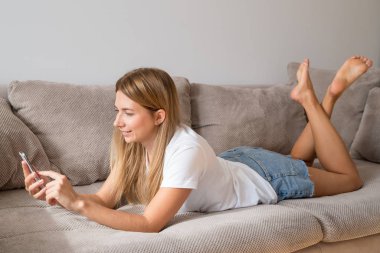 Rahat kıyafetler smartphone kullanarak ve evde kanepede yatan gülümseyen güzel kadın. 