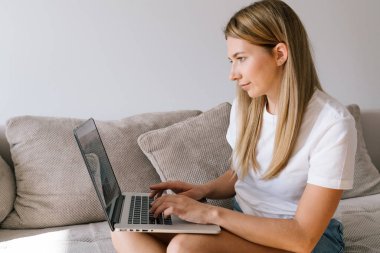 Güzel kız rahat kıyafetler içinde dizüstü kullanarak ve evde kanepede otururken gülümseyerek. İçinde tipik Internet serbest iş yapıyor klavye üzerinde beyaz T-shirt genç kadın.