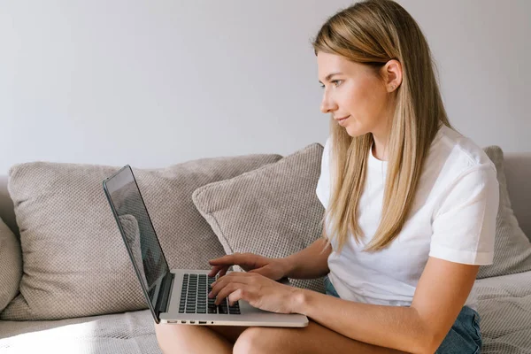 Beautiful girl in casual clothes is using laptop, and smiling while sitting on couch at home. Young woman in a white T-shirt typing on keyboard doing freelance job in internet.