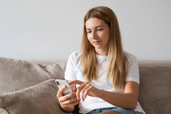 Beautiful girl in casual clothes is using a smart phone and smiling while lying on couch at home. Woman in white t-shirt browsing internet.