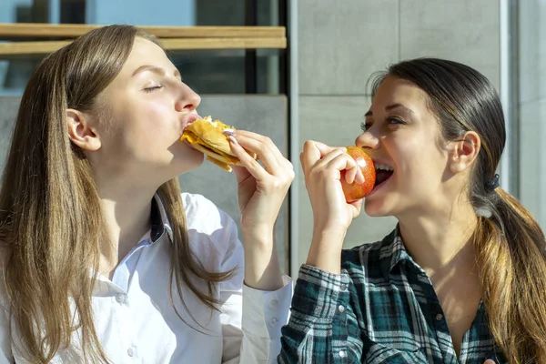 两个漂亮的女孩在咖啡馆吃一个汉堡和一个苹果 健康和垃圾食品 — 图库照片