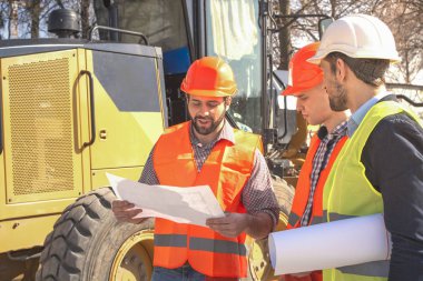 kask erkek işçiler mühendisleri buldozer ve ekskavatör yakınındaki çizimleri izliyor