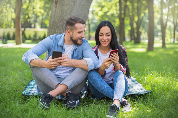 loving couple sitting on a plaid blanket in the park and use the phone, handsome guy and girl