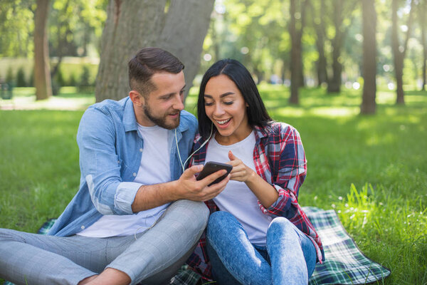 loving couple sitting on a plaid blanket in the park and use the phone, handsome guy and girl