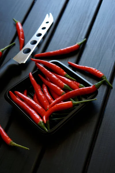 Las vainas de chile rojo y un cuchillo de verduras y queso se encuentran sobre una superficie de madera negra. Luz del día . — Foto de Stock