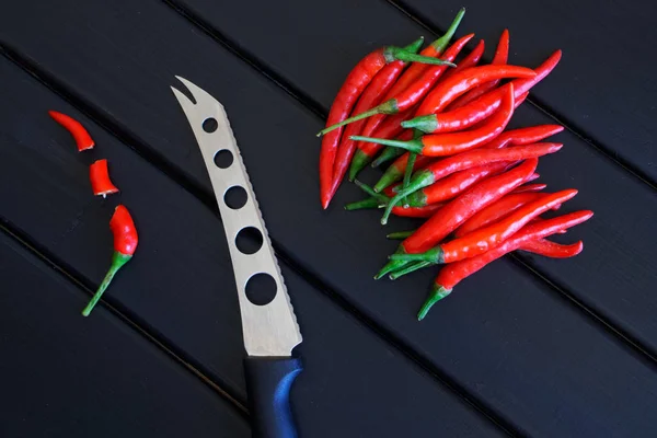 Chiles rojos cortados en rodajas y enteros junto a un cuchillo de verduras y queso yacen sobre una superficie de madera negra . — Foto de Stock