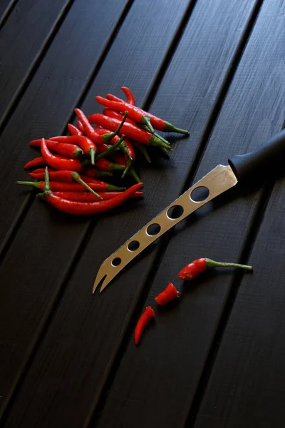 Los chiles rojos cortados en rodajas y enteros se encuentran junto a un cuchillo dentado para queso y verduras en una superficie de madera negra. Luz del día . — Foto de Stock